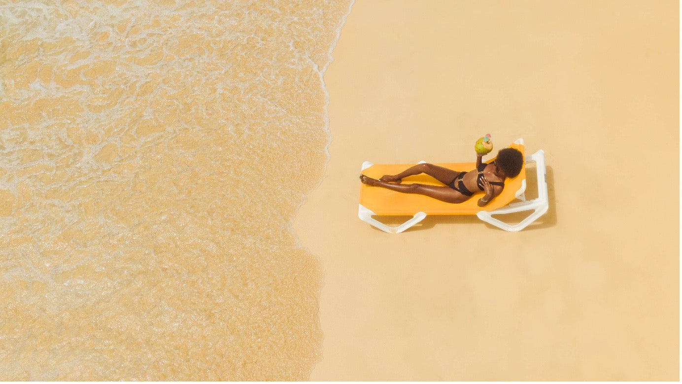 Editorial aerial shot of a woman wearing a black Aventura Swimwear bikini, relaxing on a sun lounger beside soft ocean waves on a golden beach.