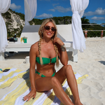 Woman in a green bikini sitting on a towel under a white canopy on a beach.