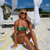 Woman in a green bikini sitting on a towel under a white canopy on a beach.