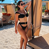 Woman in a black bikini standing on a sandy beach with lounge chairs and umbrellas in the background.